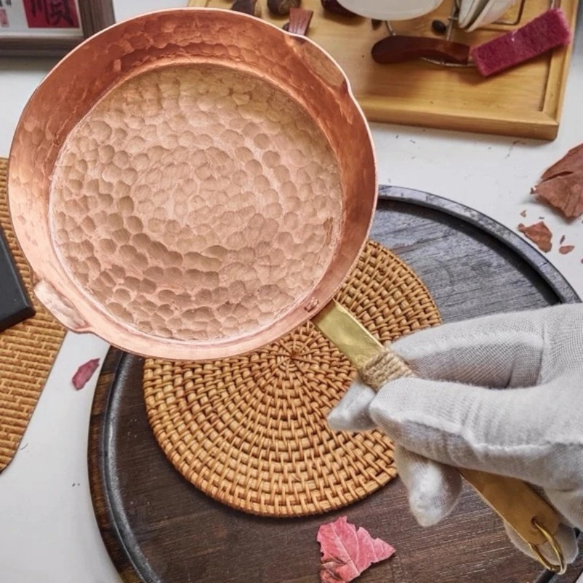 Copper Skillet close-up overhead view showing the deep hand-hammered texture inside the pure copper bowl, held by a gloved hand over a rattan trivet on a dark wooden tray