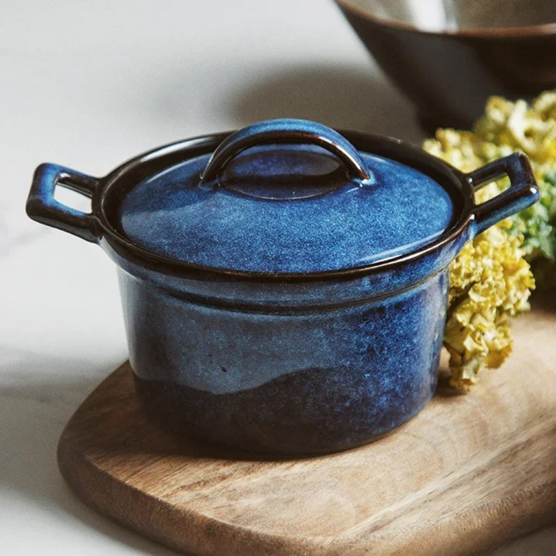 : Midnight Blue ceramic casserole with lid, styled on wooden board with dried yellow flowers beside it, dark ceramic bowl in background — Stone Vessel by CL House