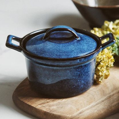 : Midnight Blue ceramic casserole with lid, styled on wooden board with dried yellow flowers beside it, dark ceramic bowl in background — Stone Vessel by CL House