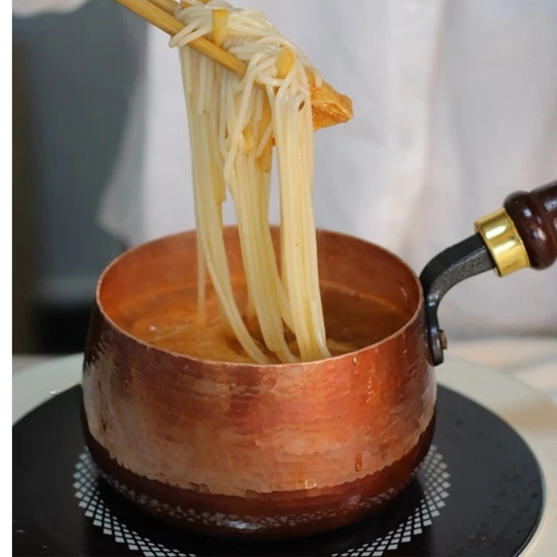  Chopsticks lifting freshly cooked noodles from the open Copper Forge saucepan on a black induction hob — hammered copper exterior glowing warm, dark wooden handle with brass fitting visible to the right, soft grey background — cooking lifestyle shot by CL House
