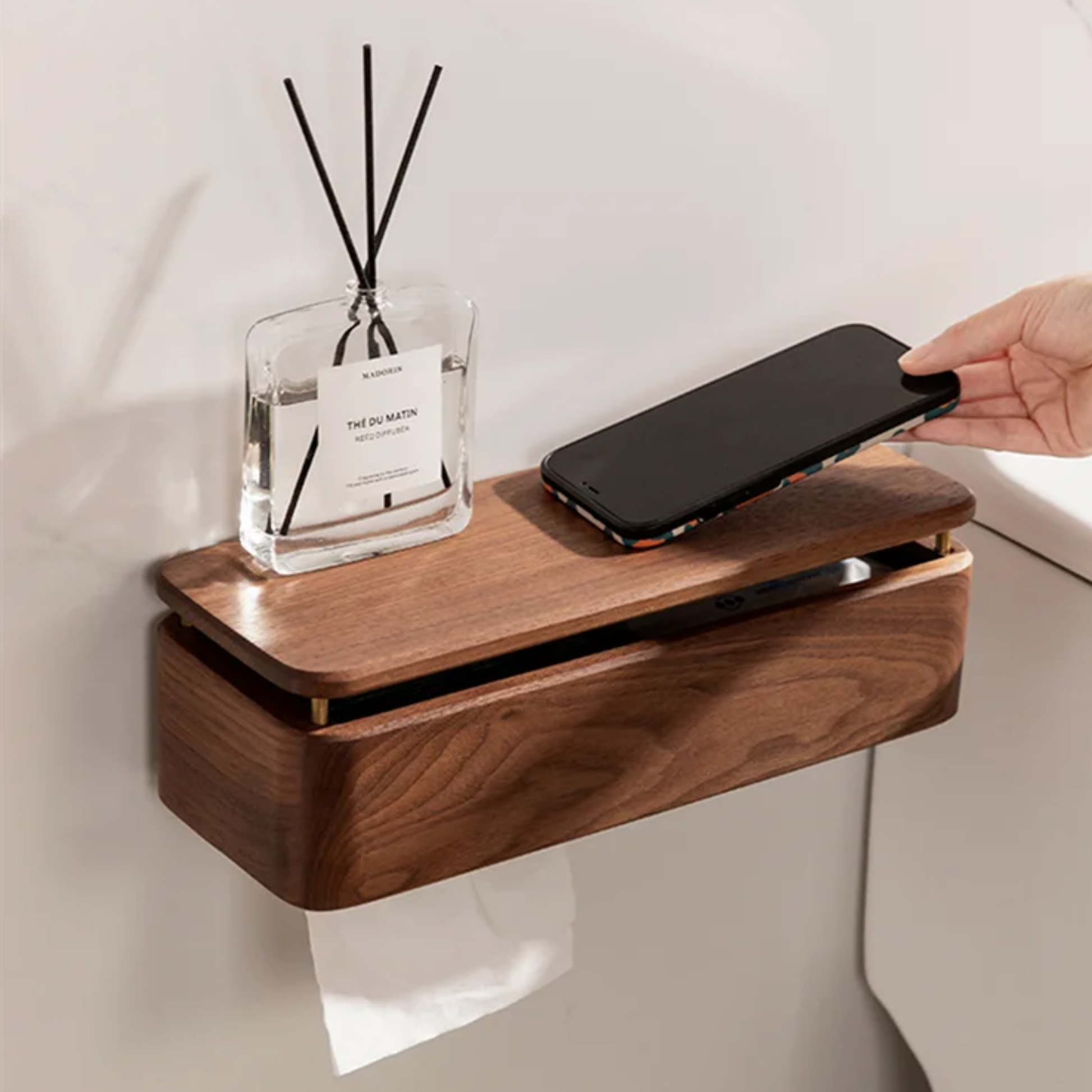 Large walnut wood wall-mounted tissue holder above toilet tank, top shelf with reed diffuser and smartphone, paper roll dispensing below — japandi bathroom lifestyle