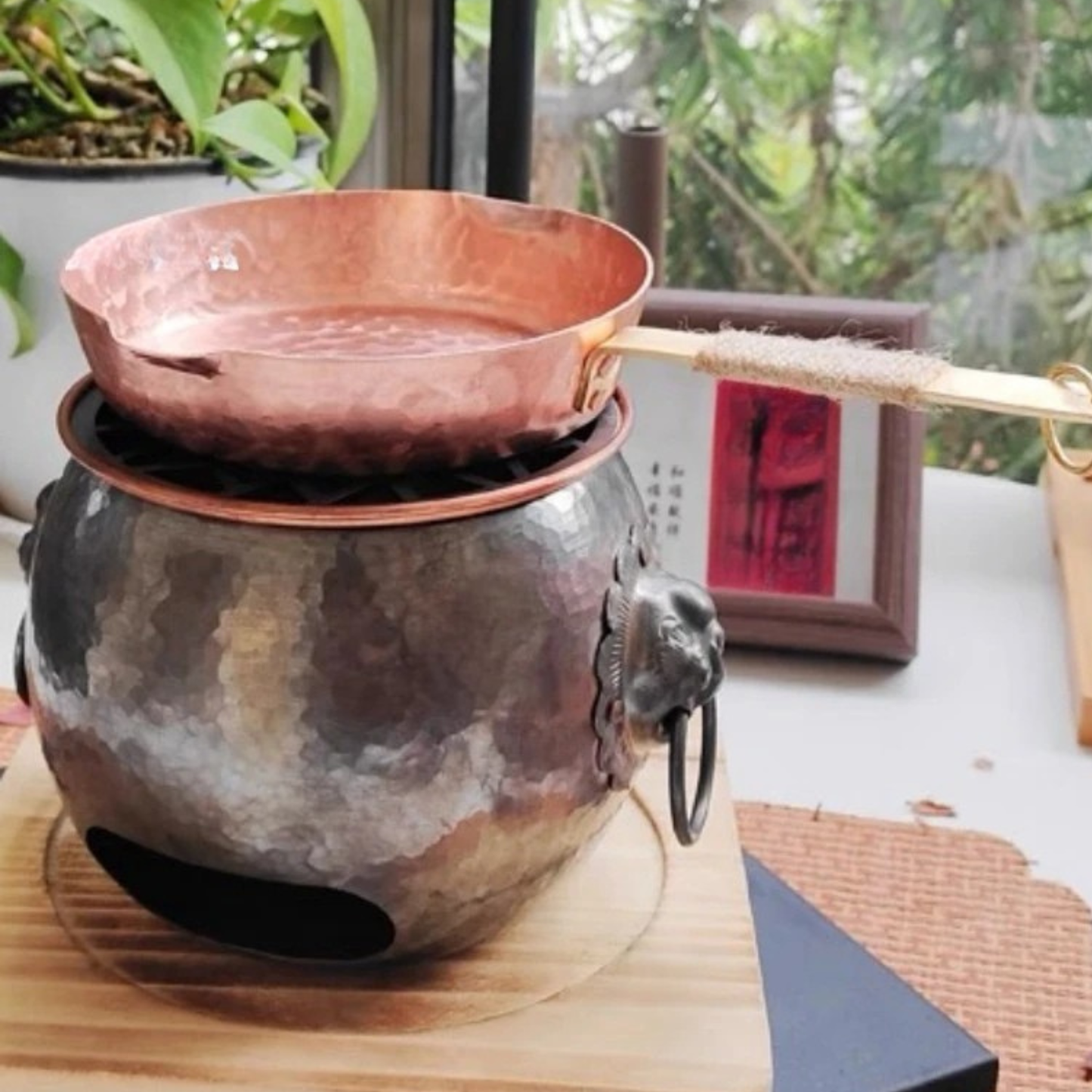 Copper Skillet pure copper frying pan resting on a traditional hammered metal charcoal stove with lion ring handles, styled on a wooden surface with indoor plants and greenery in the background