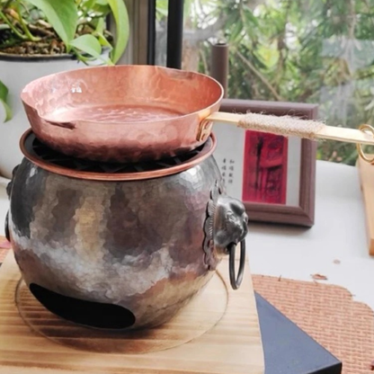 Copper Skillet pure copper frying pan resting on a traditional hammered metal charcoal stove with lion ring handles, styled on a wooden surface with indoor plants and greenery in the background