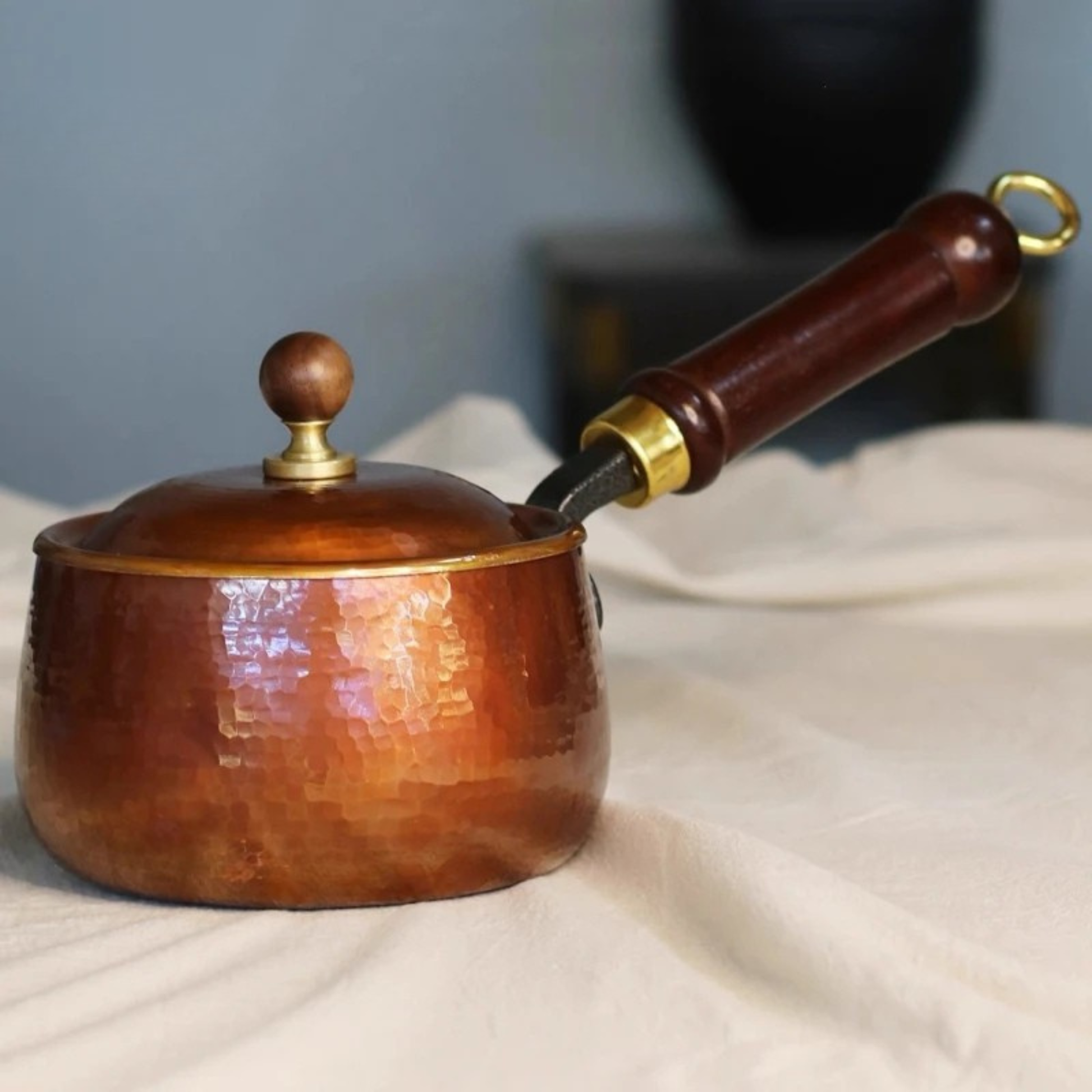  Hand-hammered pure copper saucepan with domed lid, dark wooden handle with brass fitting and hanging ring resting on white linen cloth — soft blue-grey background with dark vase in soft focus, warm copper patina highlighted by natural side light — hero lifestyle shot of the Copper Forge Saucepan by CL House