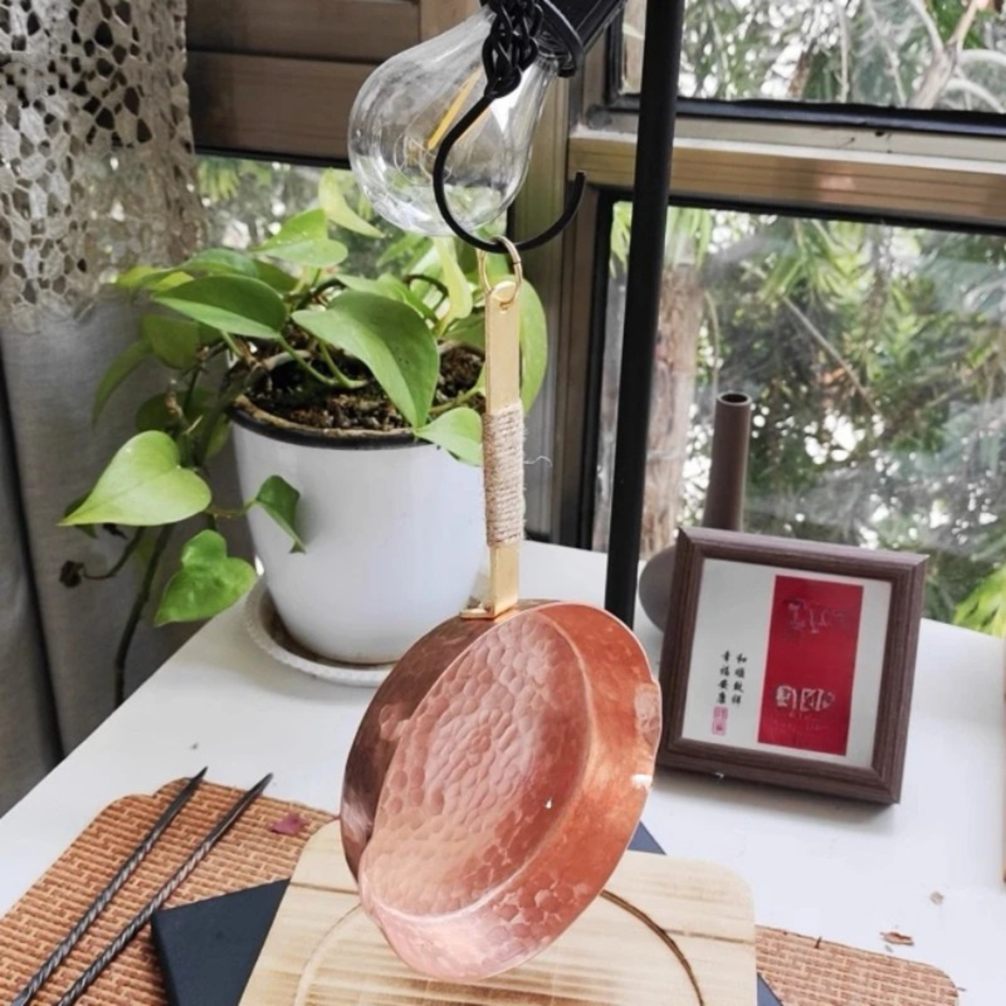 Copper Skillet pure copper frying pan hanging by its brass and jute-wrapped handle ring hook, styled on a white surface with a pothos plant and natural light from a window