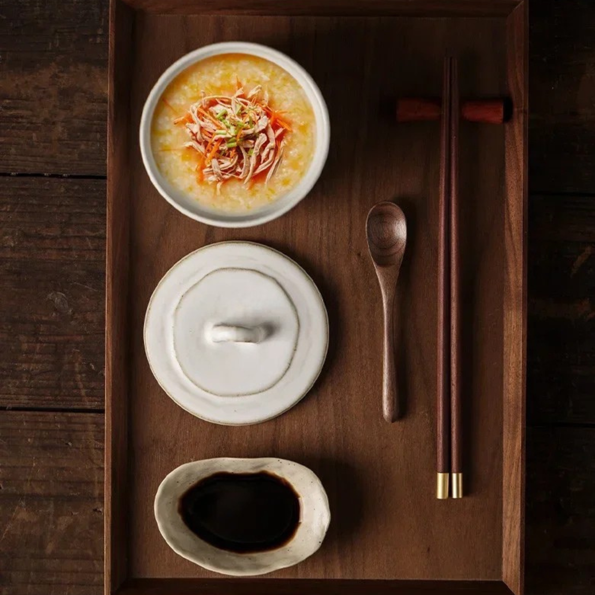 Top view of ceramic lidded bowl filled with congee topped with shredded chicken, lid and soy sauce dish arranged on a dark walnut serving tray with wooden spoon and chopsticks, japandi table setting