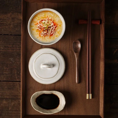 Top view of ceramic lidded bowl filled with congee topped with shredded chicken, lid and soy sauce dish arranged on a dark walnut serving tray with wooden spoon and chopsticks, japandi table setting