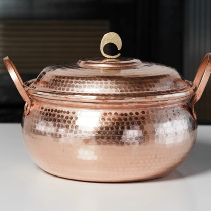 Copper Cauldron handmade pure copper stew pot with lid on a white surface against a dark background, showing the polished rose-gold hammer-textured body and brass crescent lid knob in natural kitchen light