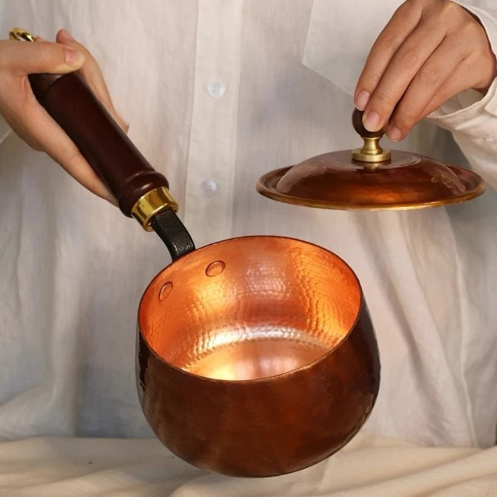 Person lifting the domed copper lid to reveal the glowing hammered pure copper interior of the Copper Forge saucepan — dark wooden handle with brass fitting held in left hand, lid raised in right hand showing wooden knob and brass detail — lifestyle interior shot by CL House