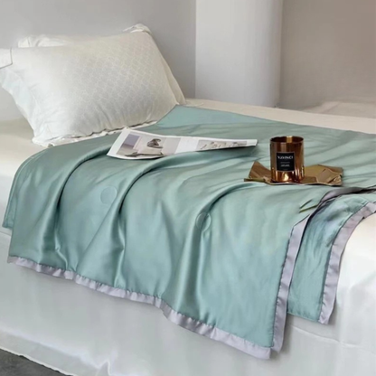 Sage Green eucalyptus lyocell cooling quilt draped over a minimalist white bed with a copper candle and open magazine as styling props, showing the silky-smooth fabric and soft lavender satin border trim