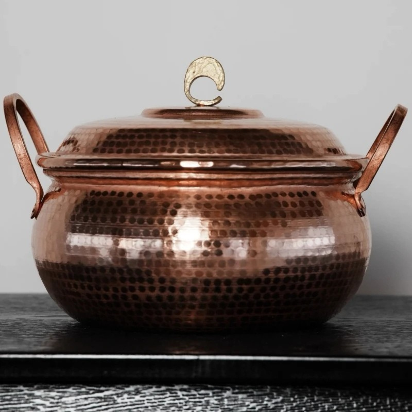 Copper Cauldron handmade pure copper stew pot with lid styled on a dark wooden surface against a grey wall, showing the deep antique copper patina, hammer-textured body and brass crescent lid knob