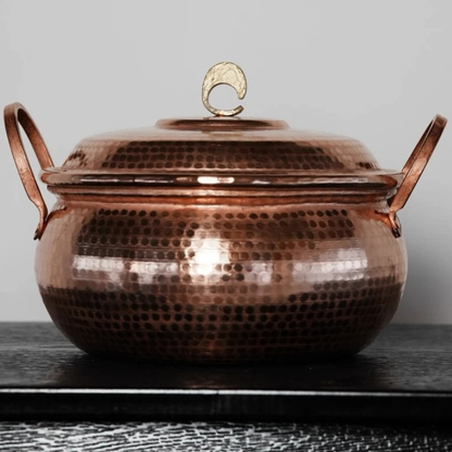 Copper Cauldron handmade pure copper stew pot with lid styled on a dark wooden surface against a grey wall, showing the deep antique copper patina, hammer-textured body and brass crescent lid knob