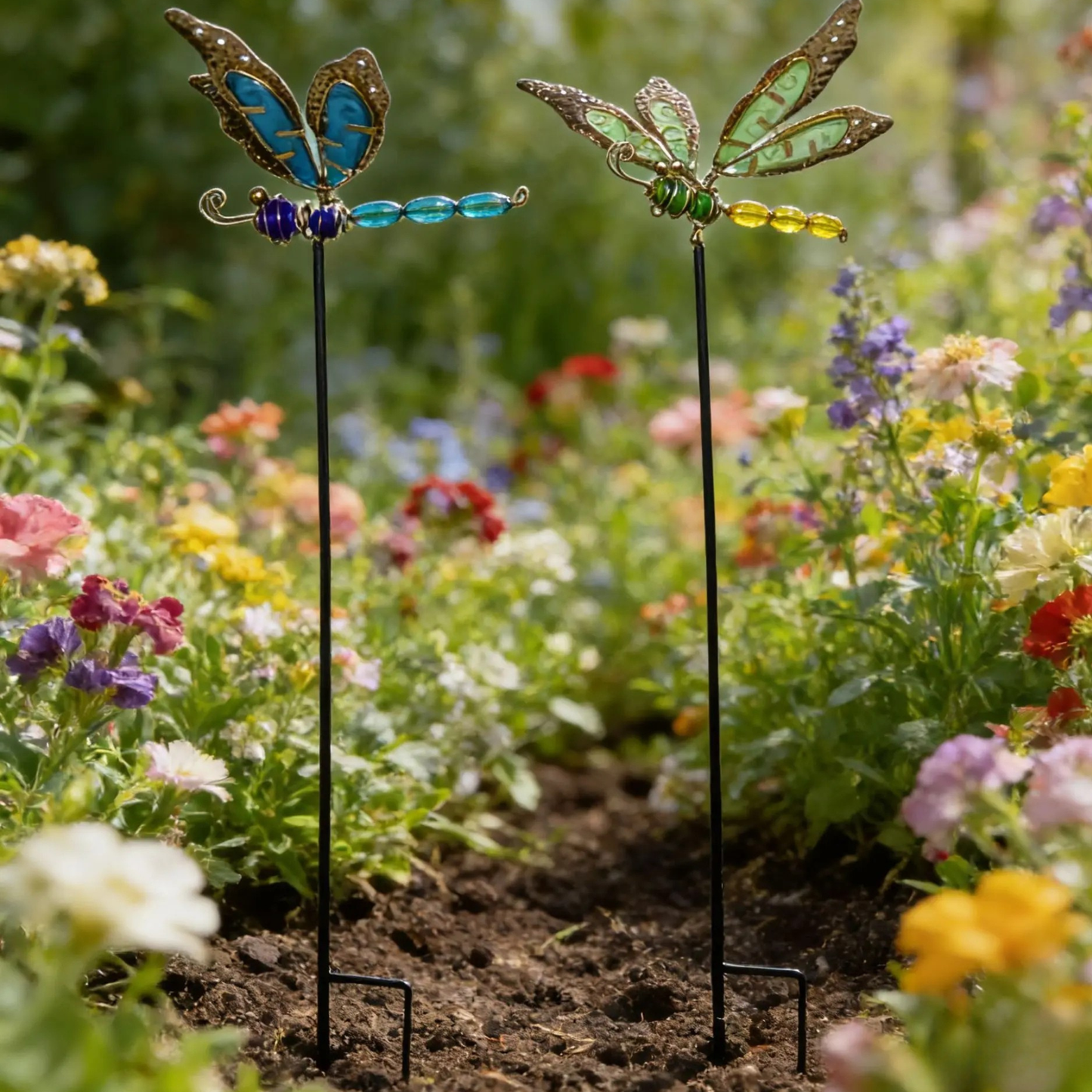 "Garden Wings" set of 2 luminous metal dragonfly garden stakes with filigree wings in blue-copper and green-gold, displayed in a colourful wildflower garden border