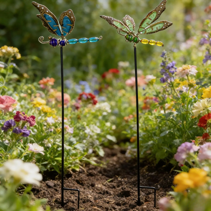 "Garden Wings" set of 2 luminous metal dragonfly garden stakes with filigree wings in blue-copper and green-gold, displayed in a colourful wildflower garden border