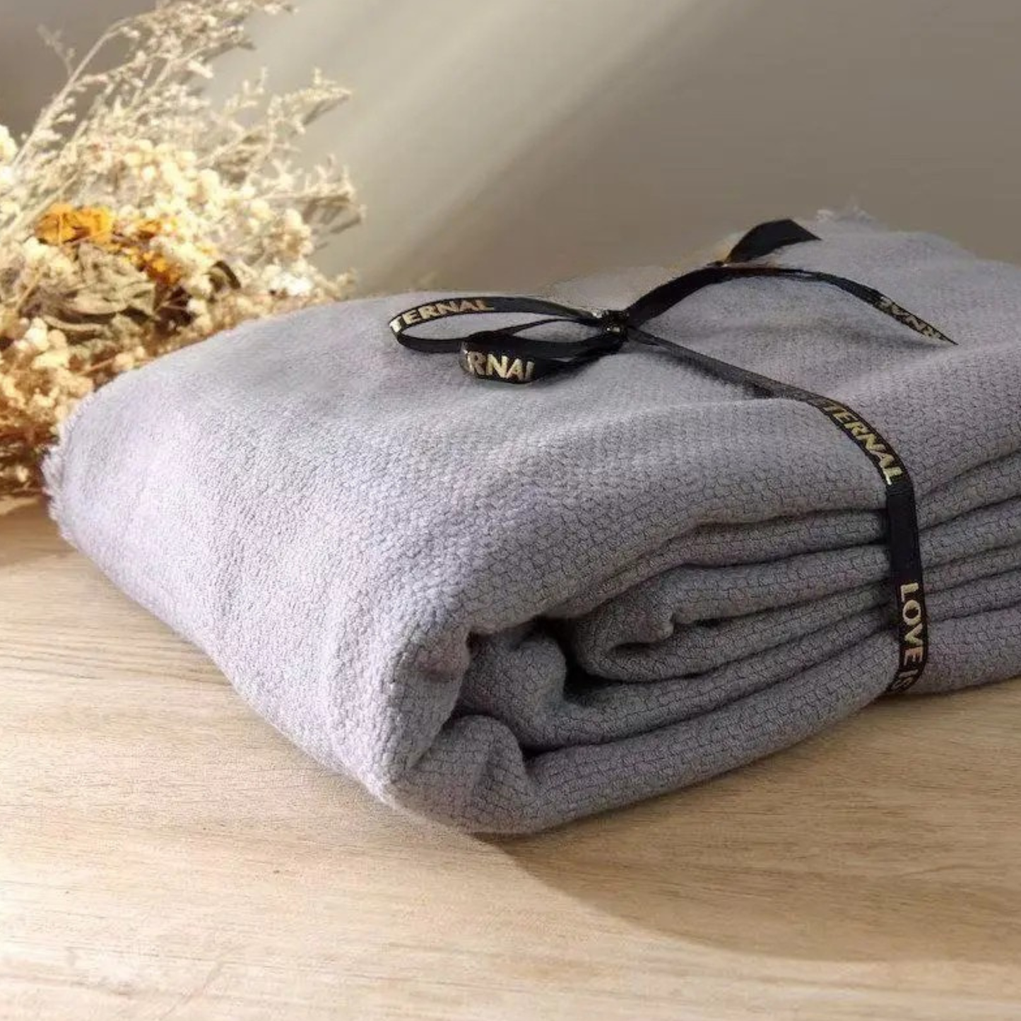  Lavender Grey cotton throw blanket folded and tied with a black ribbon bow, displayed on a wooden surface with dried botanicals in the background, showing the soft textured weave and delicate fringe trim detail