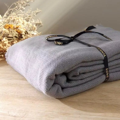  Lavender Grey cotton throw blanket folded and tied with a black ribbon bow, displayed on a wooden surface with dried botanicals in the background, showing the soft textured weave and delicate fringe trim detail