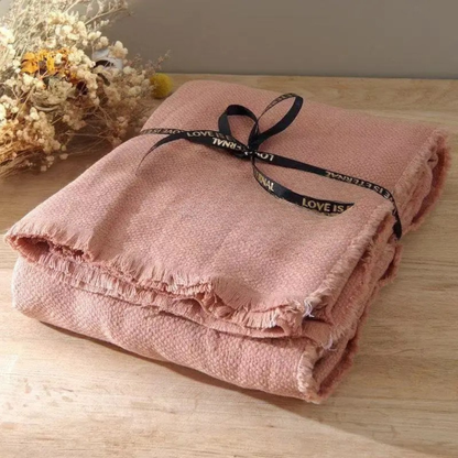 Blush Rose cotton throw blanket folded and tied with a black ribbon bow, displayed on a wooden surface with dried botanicals in the background, showing the soft textured weave and delicate fringe trim detail
