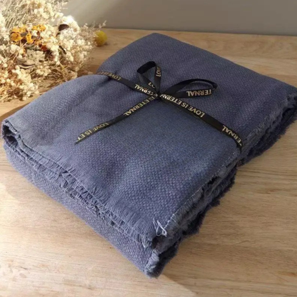 Slate Blue cotton throw blanket folded and tied with a black ribbon bow, displayed on a wooden surface with dried botanicals in the background, showing the textured waffle weave and delicate fringe trim detail