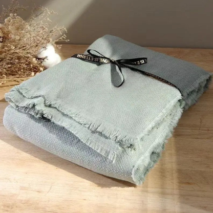 Sage Mist cotton throw blanket folded and tied with a black ribbon bow, displayed on a wooden surface with dried botanicals in the background, showing the soft textured weave and delicate fringe trim detail