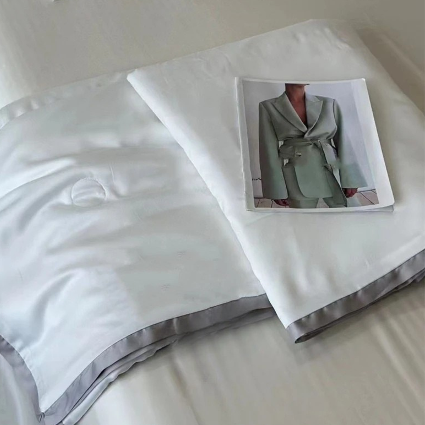  Ivory White eucalyptus lyocell cooling quilt folded and displayed against a white background with a fashion editorial print as a styling prop, showing the silky-smooth fabric and soft grey satin border trim