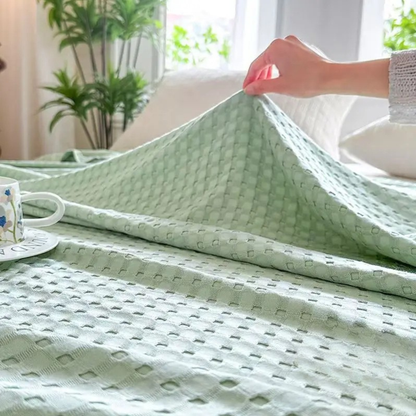 "Hand lifting 100% Bamboo Fibre waffle-texture cooling blanket in Sage Mist — showing the soft double-sided weave and lightweight feel, breathable bamboo summer blanket styled on a bed with a floral cup"