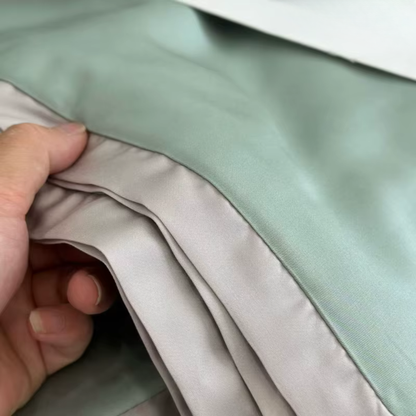 Close-up of a hand lifting the soft grey satin border trim of the Sage Green eucalyptus lyocell cooling quilt, showing the lightweight silky fabric and refined edge finishing detail