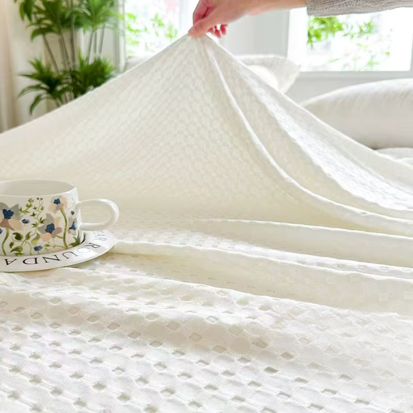 "Hand lifting 100% Bamboo Fibre waffle-texture cooling blanket in Ivory White — showing the soft double-sided weave and lightweight feel, breathable bamboo summer blanket styled on a bed with a floral cup"