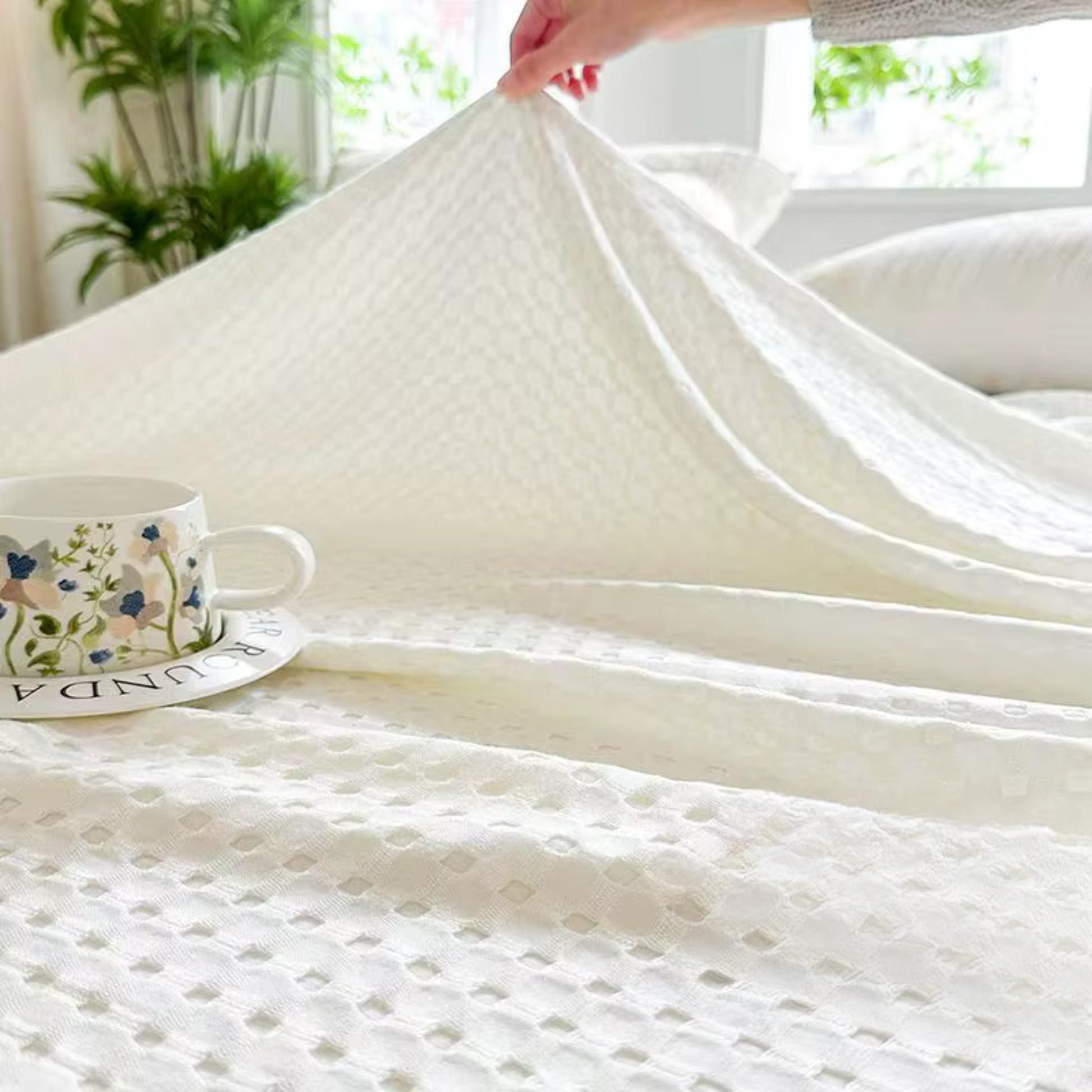 "Hand lifting 100% Bamboo Fibre waffle-texture cooling blanket in Ivory White — showing the soft double-sided weave and lightweight feel, breathable bamboo summer blanket styled on a bed with a floral cup"