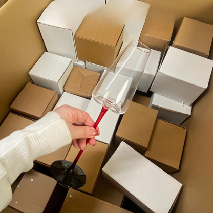 Hand holding a "Stem & Base" crystal champagne flute in Crimson Stem variant over its packaging boxes — CL House Drinkware collection