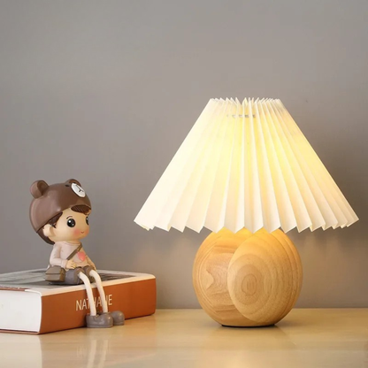  "Pleat & Wood" pleated table lamp with off white shade and round wood sphere base glowing warmly, styled on a wooden desk with a decorative figurine