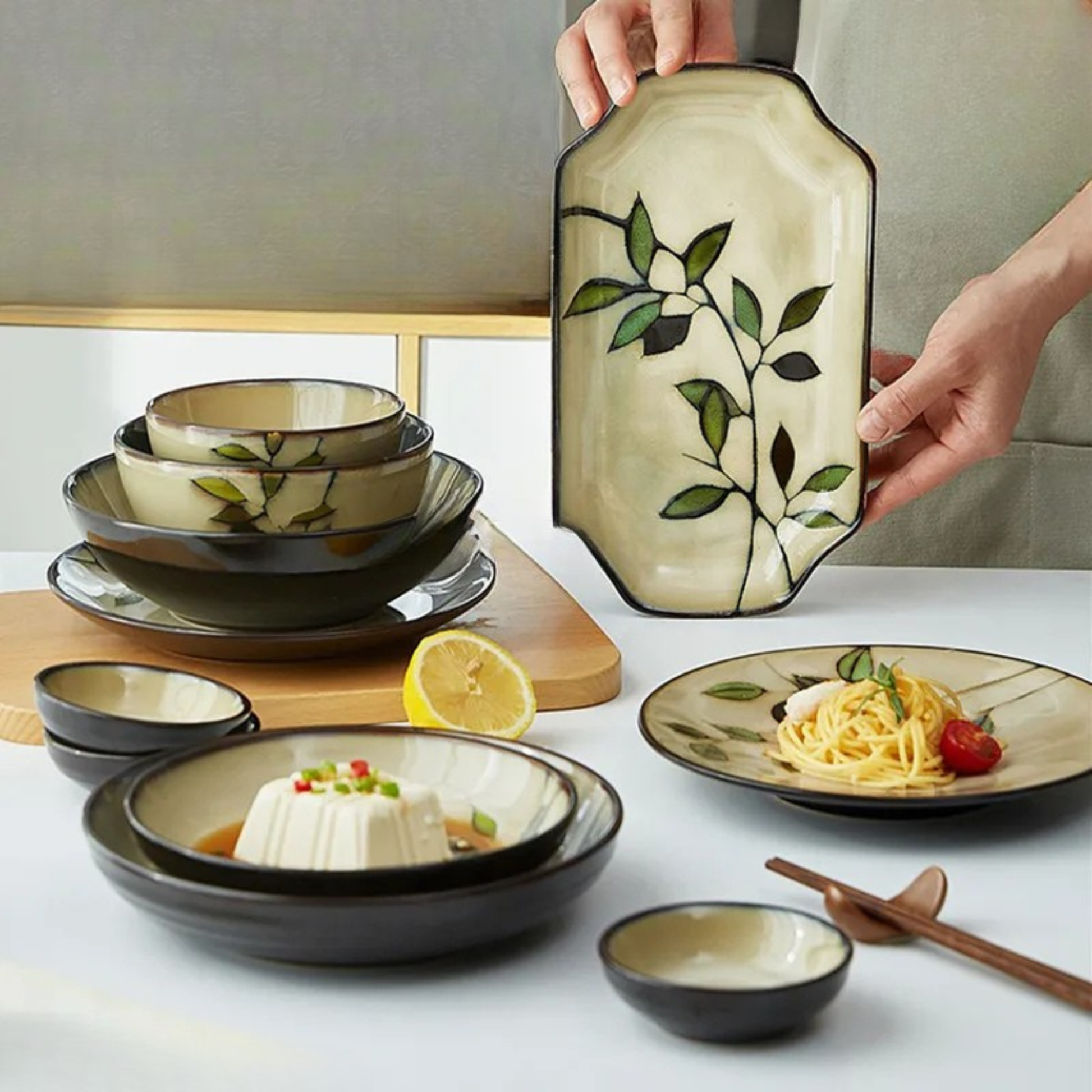 "Painted Leaf" ceramic tableware collection styled on a dining table, including soup bowls, dinner plate with pasta, rectangular serving tray held by hand, hand-painted leaf motif in ivory and espresso glaze, japandi dining setting