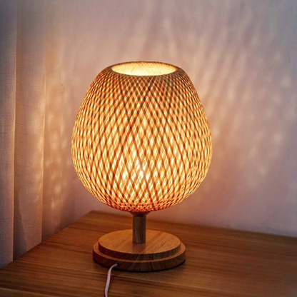 Woven bamboo table lamp illuminated on a wooden nightstand, casting warm light patterns on the wall — "Woven Glow" Small 23 × 26cm by CL House