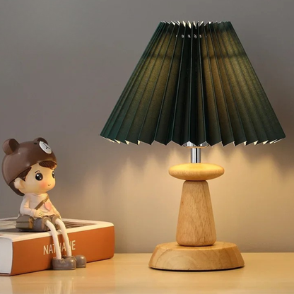 "Pleat & Wood" pleated table lamp with forest green shade and spool-shaped wood base, warm ambient light on a wooden desk with a decorative figurine