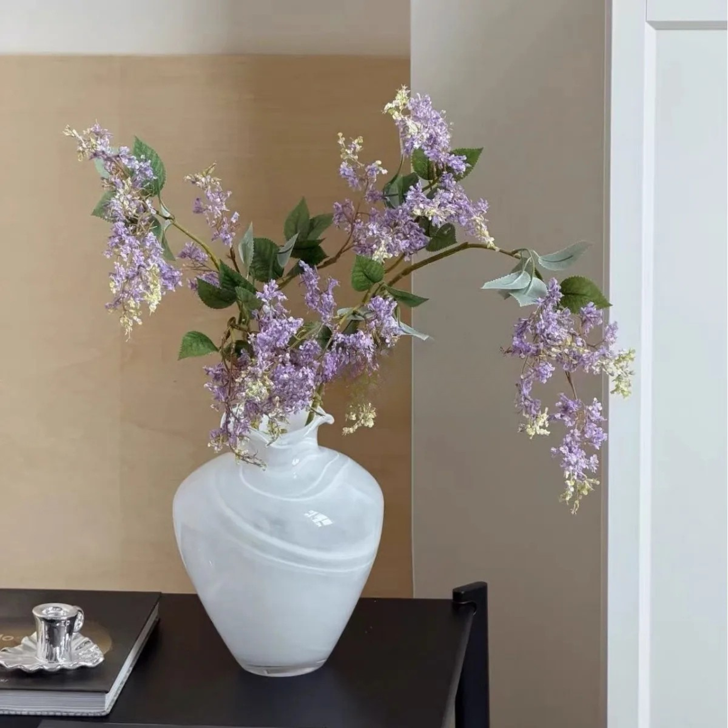 "Petal Wave" handblown glass vase in Cloud Drift filled with cascading purple lilac branches — white swirl glass, styled on dark console table with silver candle holder, warm neutral background