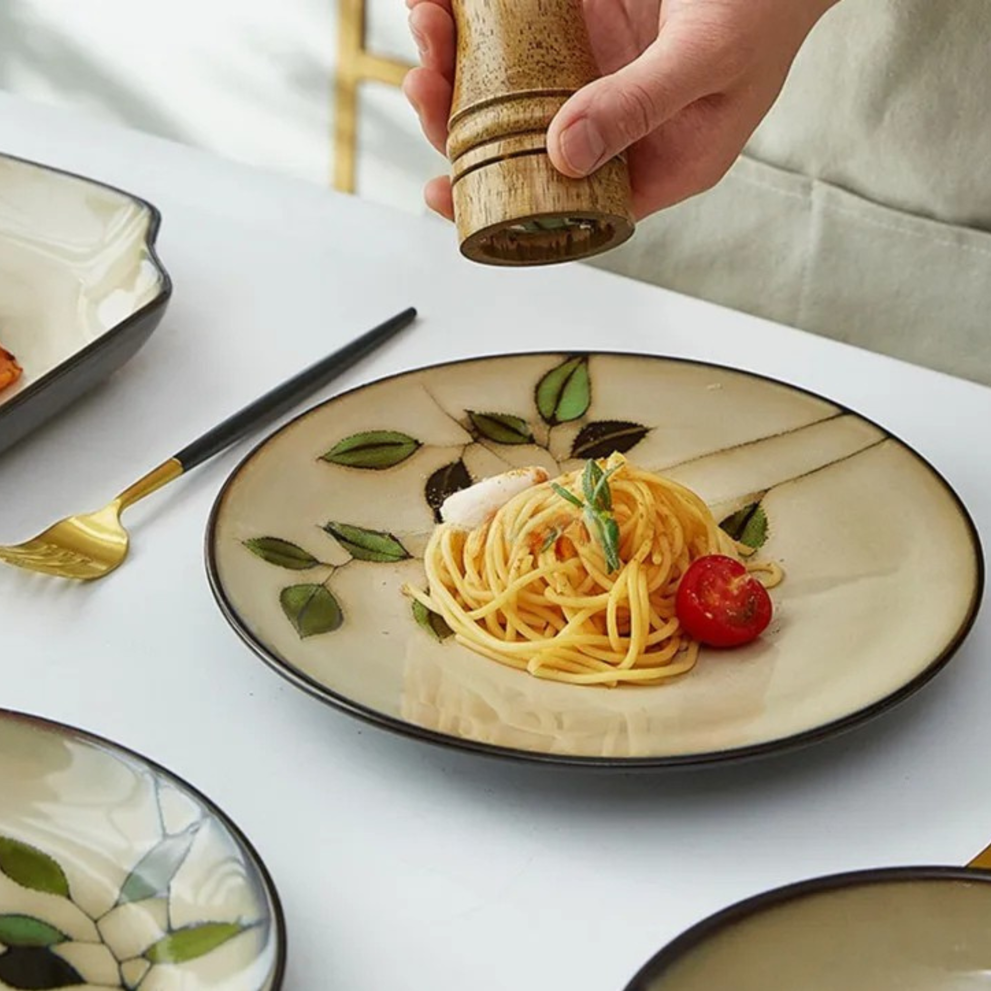  "Painted Leaf" ceramic dinner plate styled with pasta and cherry tomato, hand holding a wooden pepper mill, japandi tableware in a dining table setting