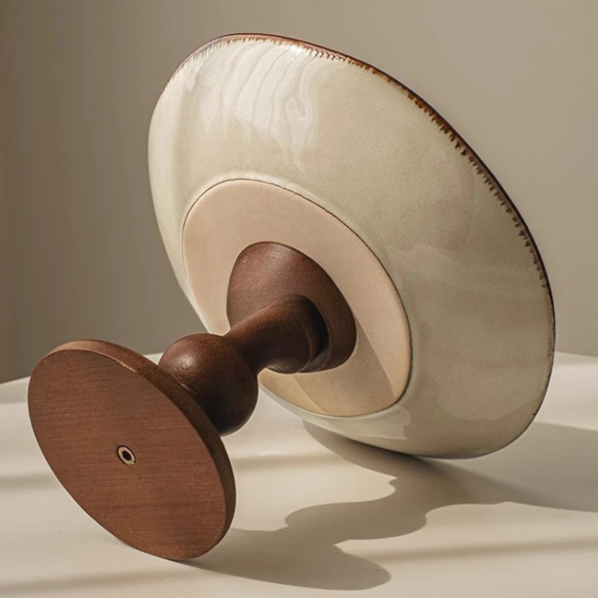 "Blue Botanics" ceramic pedestal serving stand seen from below, revealing the solid walnut wood turned base and the clean ivory ceramic underside — detail shot