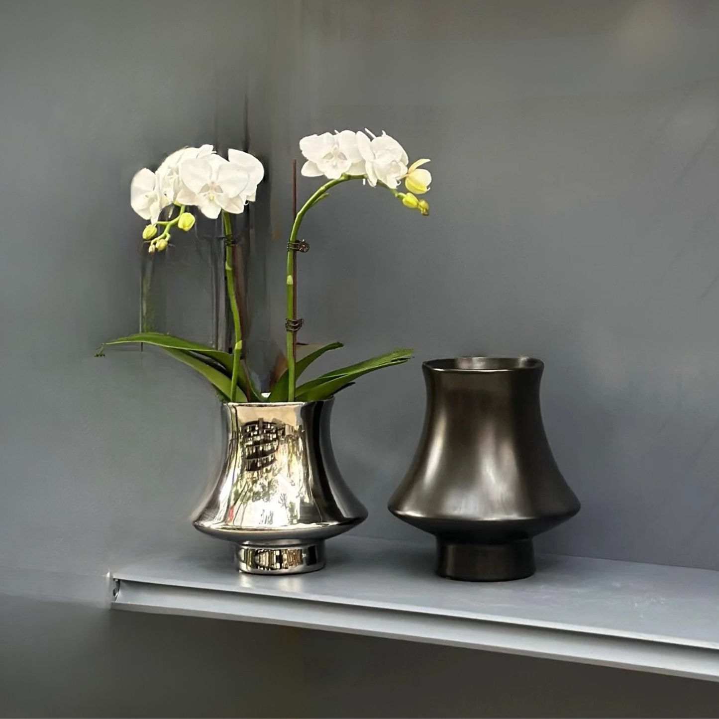Bell Tower metallic ceramic vases in Silver and Gunmetal finishes styled with white orchid stems on grey shelf