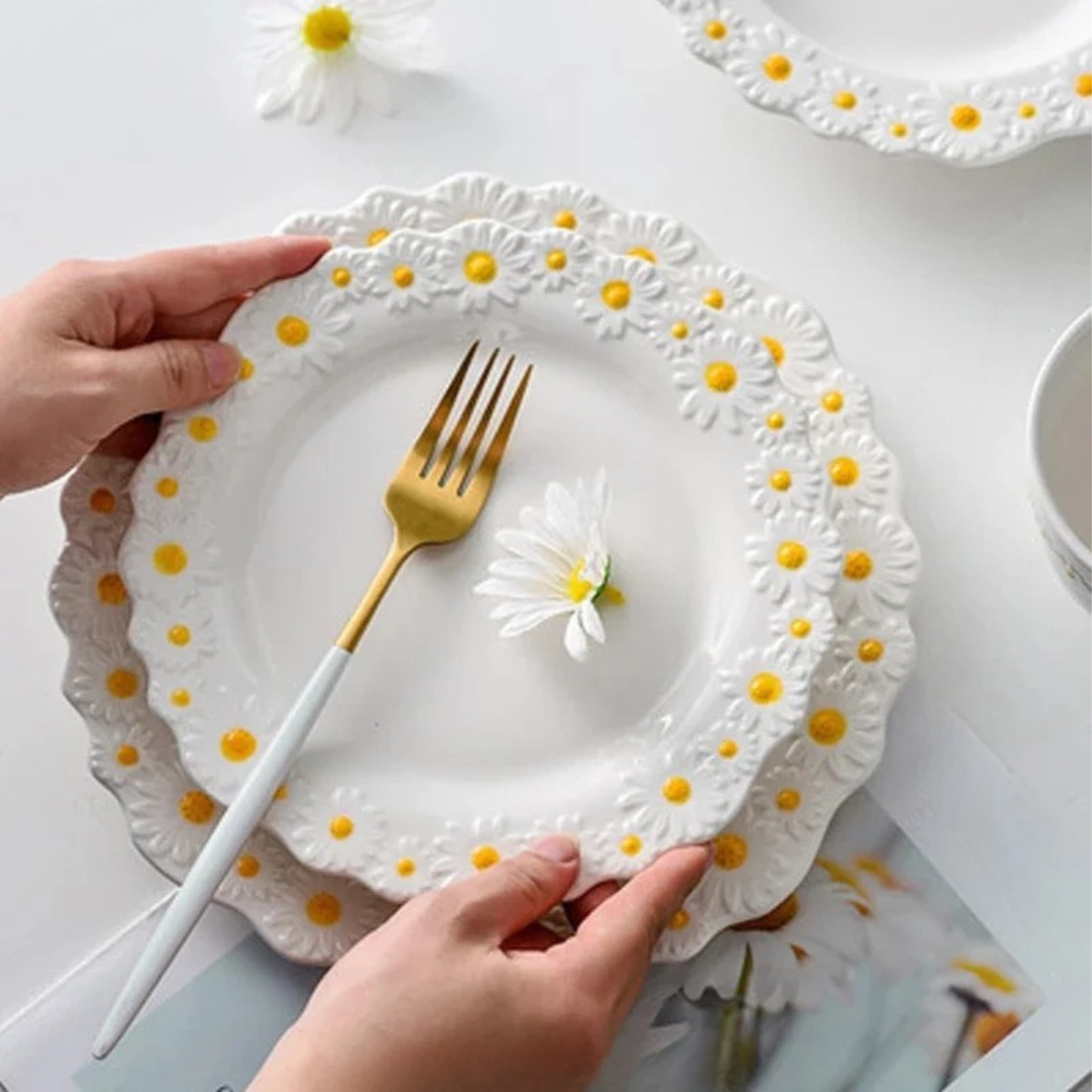 "Daisy Bloom" ceramic dinner plate — top view styled with a gold fork and fresh daisy flower on white table surface, showing stacked big and small plates with raised daisy relief border — lifestyle shot