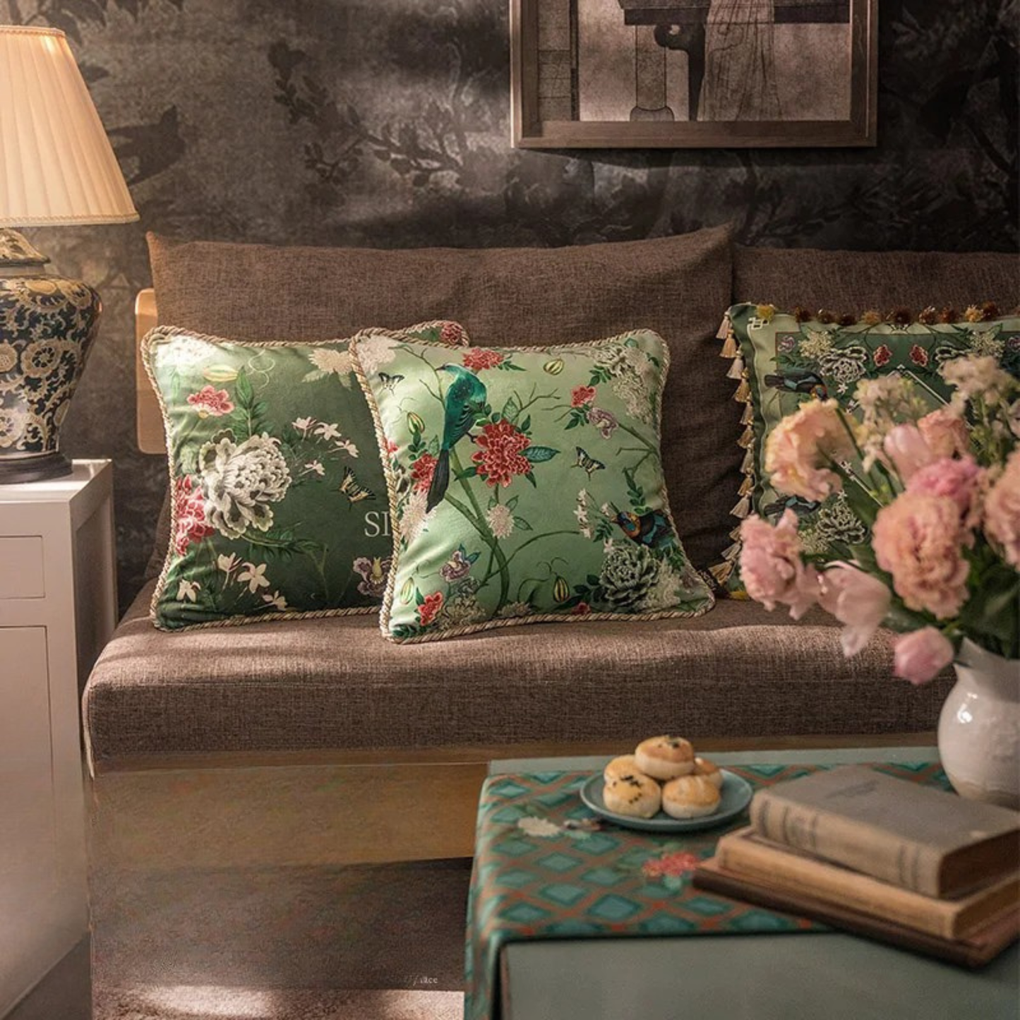  "Garden Silk" velvet cushion covers styled on a linen wooden sofa — Silken Blooming and Springing designs in deep and sage green, chinoiserie botanical print, with braided piping and tassel trim variants, Chinese porcelain lamp and fresh pink peonies