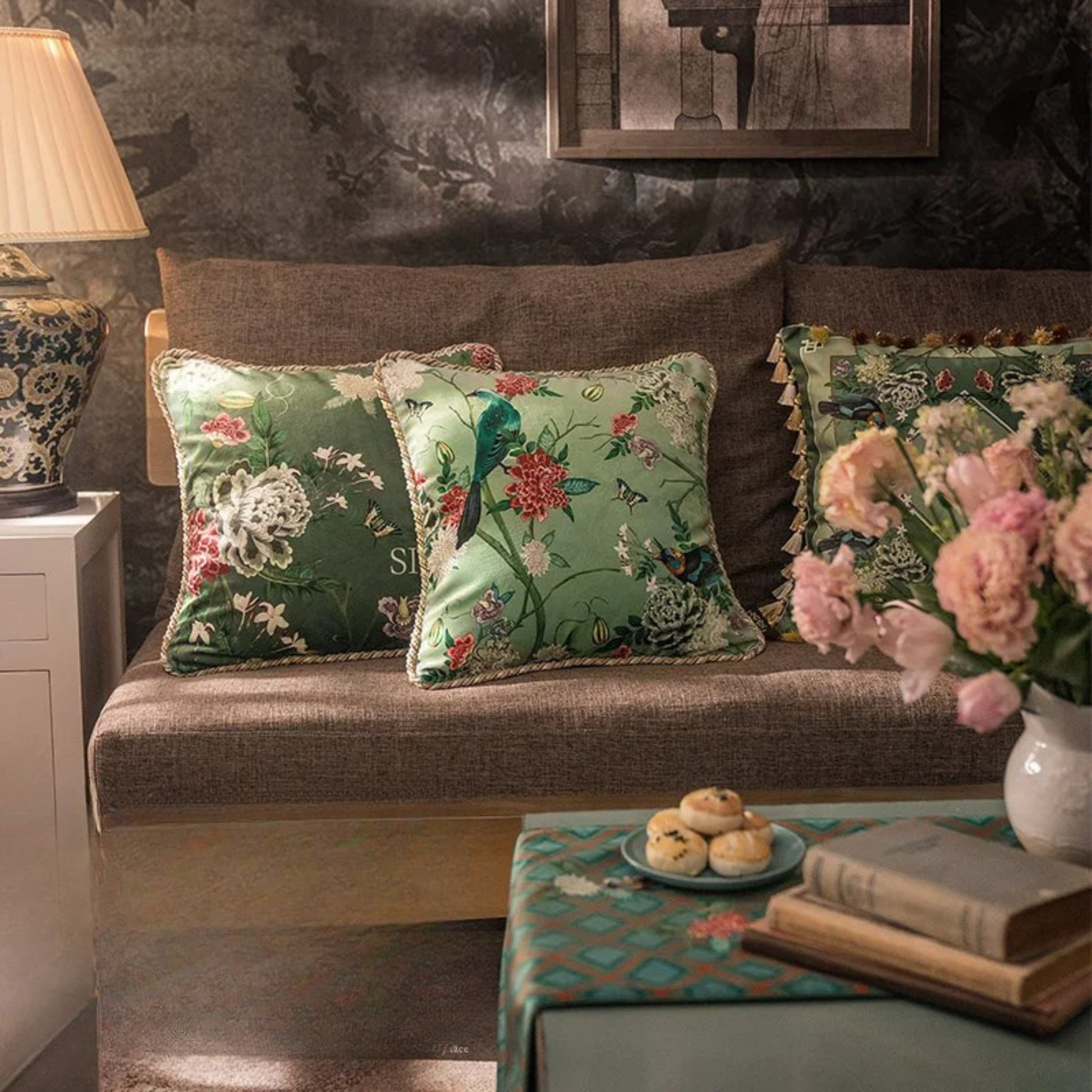  "Garden Silk" velvet cushion covers styled on a linen wooden sofa — Silken Blooming and Springing designs in deep and sage green, chinoiserie botanical print, with braided piping and tassel trim variants, Chinese porcelain lamp and fresh pink peonies