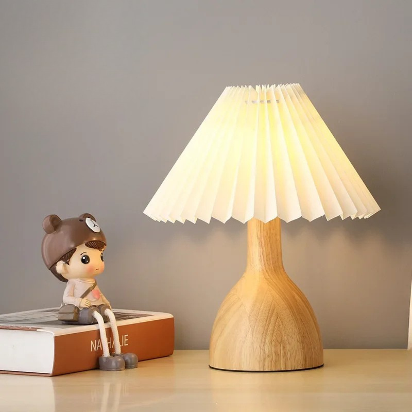 "Pleat & Wood" pleated table lamp with off white shade and vase-shaped wood base, warm glow on a wooden desk with a decorative figurine