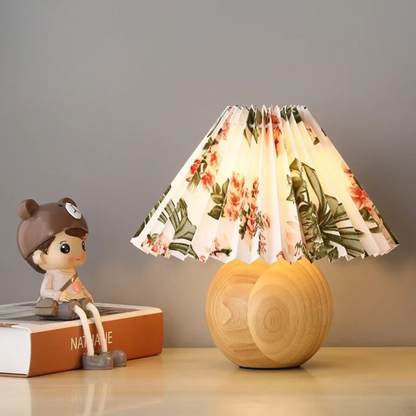 "Pleat & Wood" pleated table lamp with floral shade and round wood sphere base, styled on a bedside table with a decorative figurine